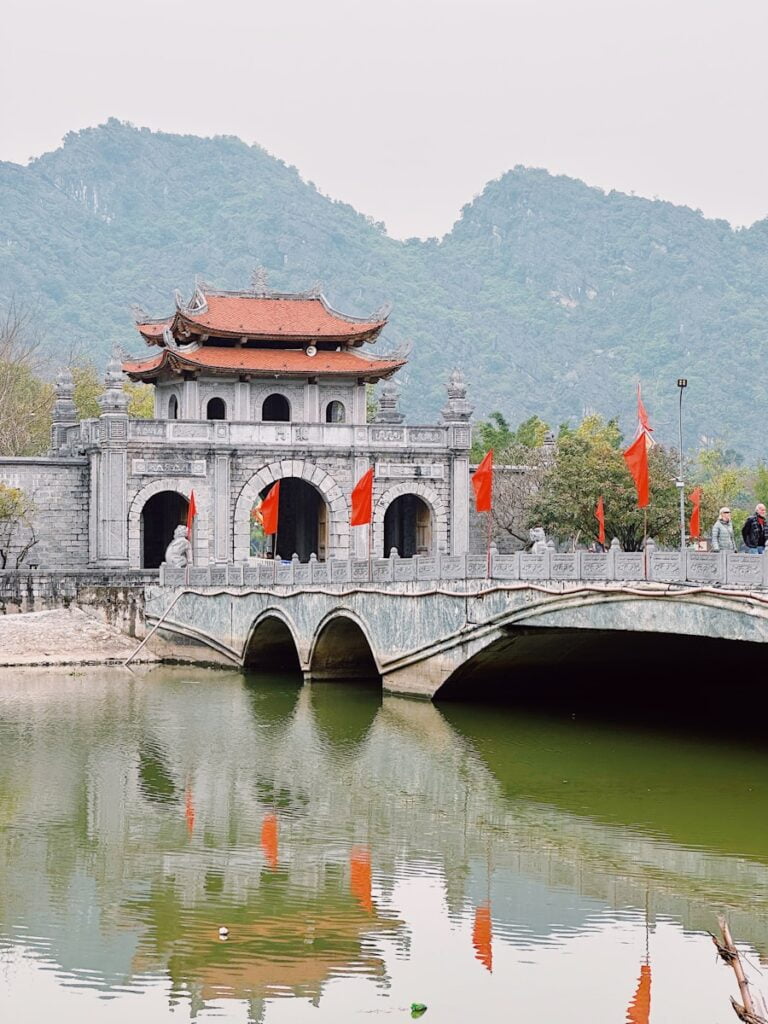 A bridge spans a river, with pedestrians crossing and a building visible in the background during a Ninh Binh day trip.